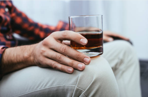 Close-up of a person holding a glass of dark beverage (likely whiskey) resting on their knee in a casual setting.