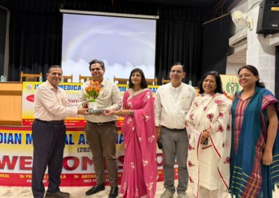 Six people stand on a stage, with one man handing a potted flower to another as a group photo during a ceremony, with banners in the background.