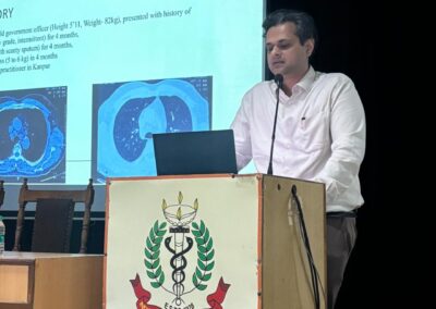Male presenter at a podium giving a medical lecture, with projection slides showing medical images behind him.