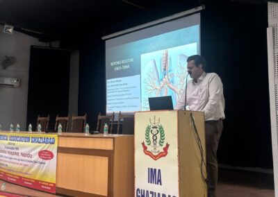 Speaker at a podium giving a presentation on stage, projector screen showing a medical slide titled 'Beyond Routine' with 'IMA GHAZIABAD' banners nearby.