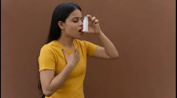 Asthma - drmanumadan.com Woman in a yellow shirt holds a small bottle to her mouth with her right hand while touching her chest with her left hand, against a brown wall.