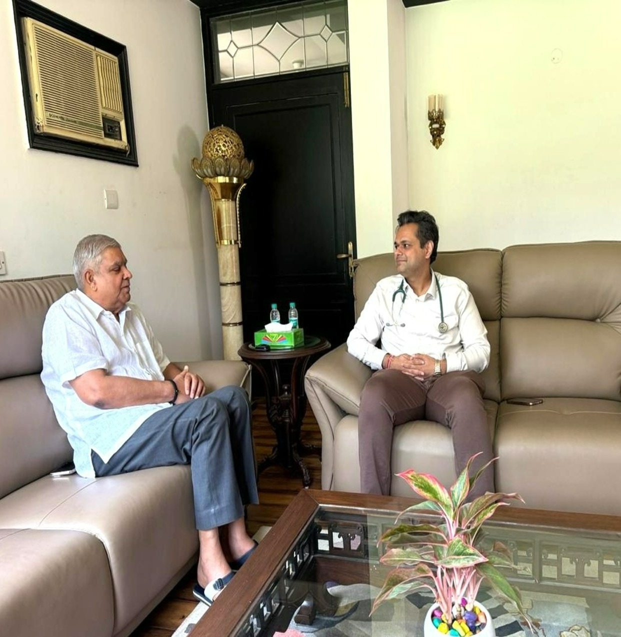 WhatsApp Image 2026-10-06 at 60504 PM - drmanumadan.com Two men sit on beige couches in a living room, one older in a white shirt chatting with a doctor wearing a stethoscope.