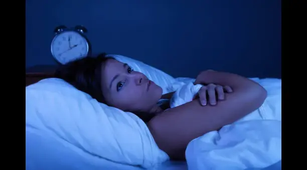 sleep-disorders - drmanumadan.com Woman awake in bed at night, lying on her side under white blankets with an analog alarm clock on the nightstand in a blue-lit room.
